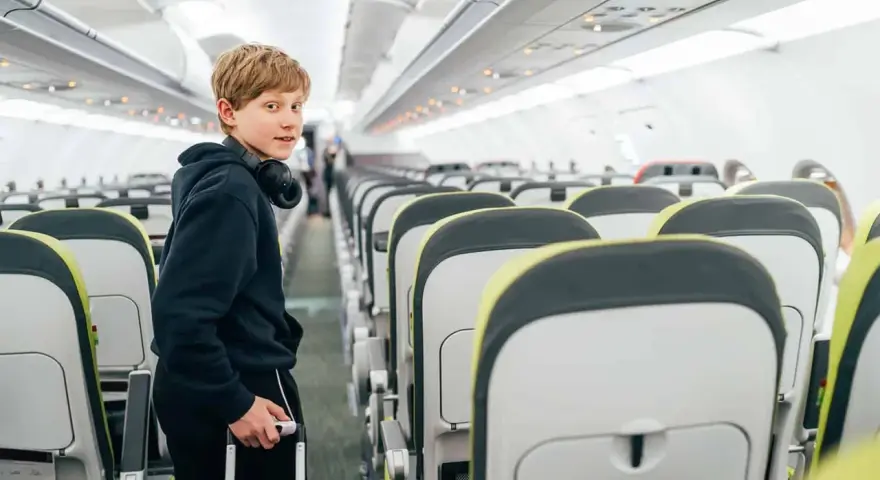 Chłopiec z słuchawkami na szyi i walizką w ręku stoi w przejściu samolotu. Od ilu lat można latać samemu? Ryanair pozwala na samodzielne podróże od 12 roku życia.