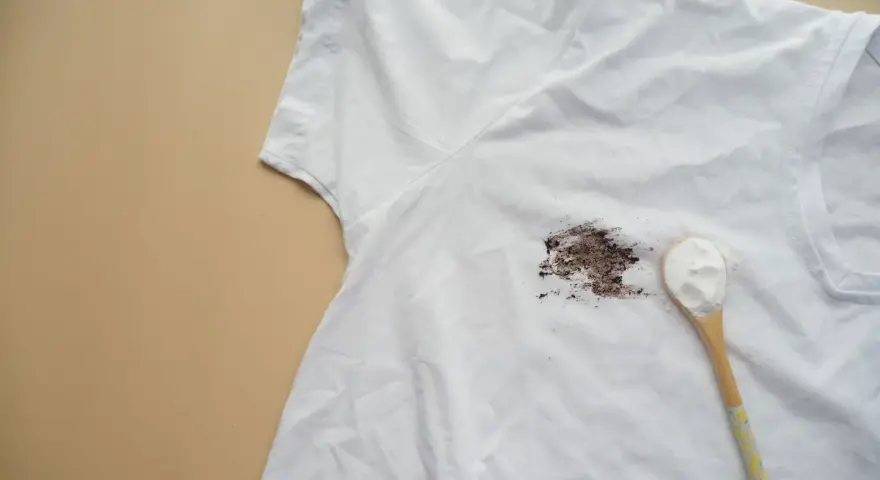 Tache sur t-shirt blanc après lavage, avec une cuillère de poudre blanche à côté.