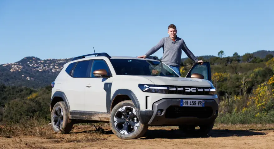 Mężczyzna opiera się o maskę jasnoszarego Dacia Duster, hybrydy z fabrycznym LPG, na tle wzgórz.