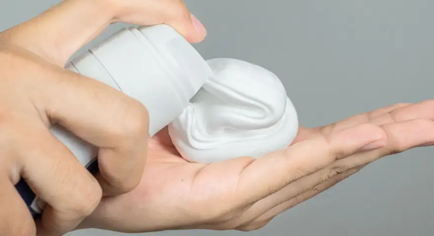 Mano aplicando gel o espuma de afeitar de un envase blanco a la palma.