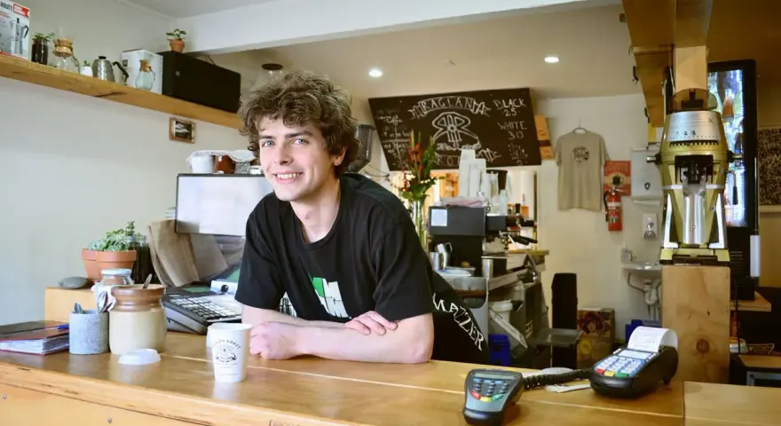 Ein junger Mann mit lockigen Haaren lächelt hinter der Theke eines Cafés. Er scheint seine **zwei Werkstudentenjobs** zu genießen, während er Kaffee zubereitet.