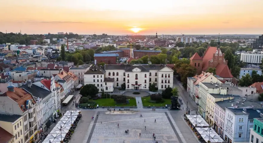 Zachód słońca nad rynkiem, gdzie widać piękną pierzeję kamienic i ratusz. Ludzie spacerują po placu, ciesząc się ciepłym wieczorem.