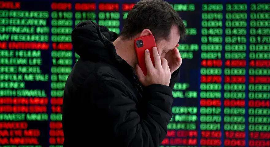 Hombre preocupado habla por teléfono frente a pantalla de bolsa de valores de Nueva York, con números rojos y verdes indicando un posible crack.