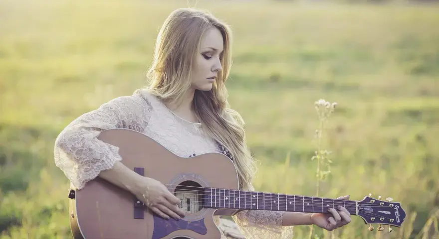 Blondynka w koronkowej sukience gra na gitarze w słonecznym polu. To jak remaster starych wspomnień, pełen ciepła i nostalgii.