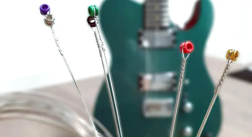 Vier Gitarrensaiten mit farbigen Enden (lila, grün, rot, gold) liegen vor einer grünen E-Gitarre. Die besten E-Gitarren-Saiten für deinen Sound.