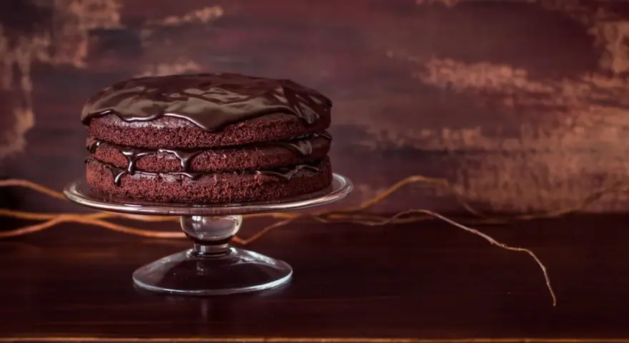 Torta al cioccolato a tre strati, lucida e invitante, su un'alzata di vetro. Gocce di glassa scivolano sui bordi.
