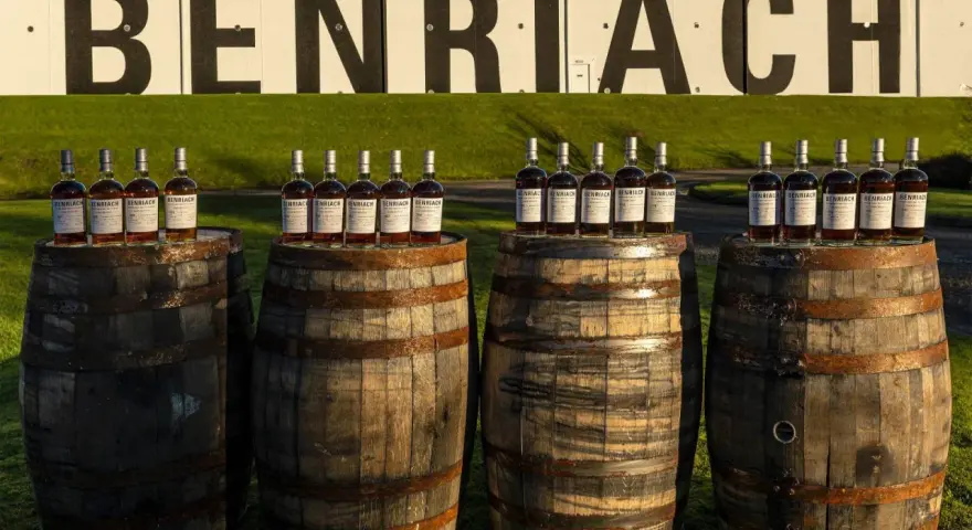 Butelki whisky Benriach stoją na beczkach, które czekają na dojrzewanie w pomieszczeniu.