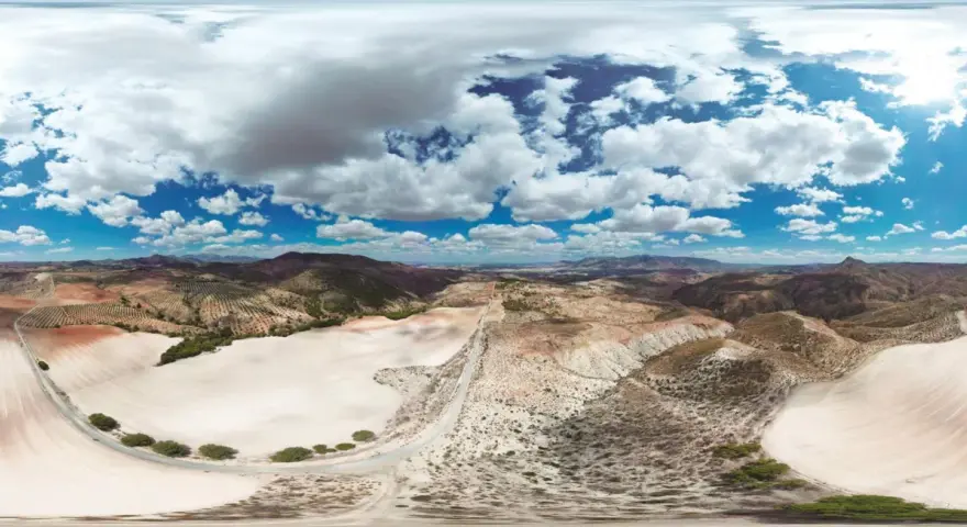 Paisaje árido con colinas, campos cultivados y un camino serpenteante. El cielo azul con nubes blancas contrasta con el desierto de larva.