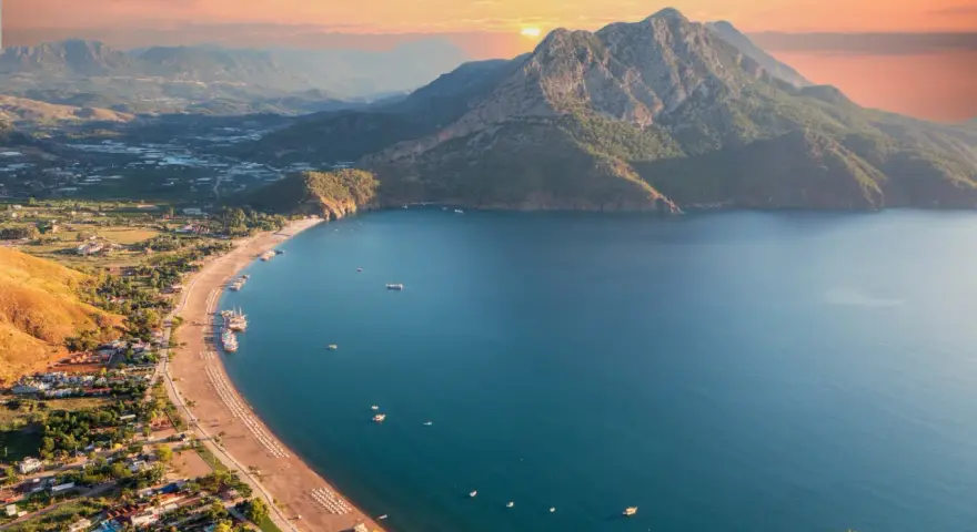 Malownicze wybrzeże Turcji o zachodzie słońca. Góry schodzą do turkusowego morza, a plaża z leżakami i łodziami zaprasza do odpoczynku.