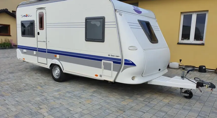 Biała przyczepa kempingowa Hobby 460 UFE "de luxe easy" stoi na kostce brukowej przed żółtym budynkiem.