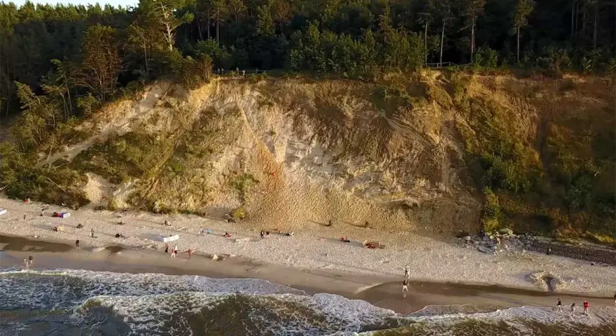 Plaża u stóp najwyższego klifu w Polsce. Ludzie spacerują po piasku, fale obmywają brzeg.