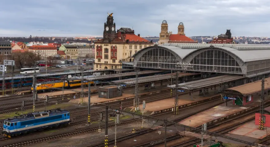 Widok na dworzec kolejowy w Pradze z pociągami na torach i budynkami w tle.