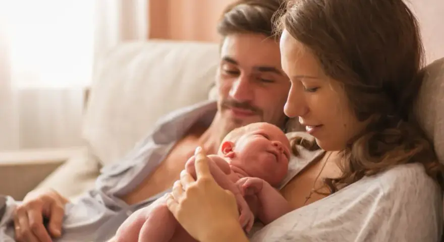 Mama tuli noworodka, tata patrzy z miłością. To piękny obraz opieki na żone po porodzie, pełen czułości i wsparcia.