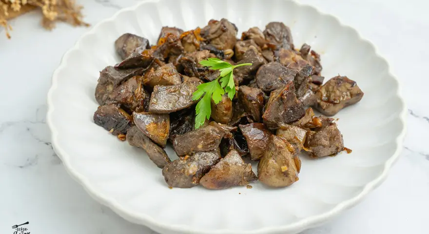 Plato de asadura de cordero salteada, con trozos oscuros y jugosos, adornado con una ramita de perejil fresco.
