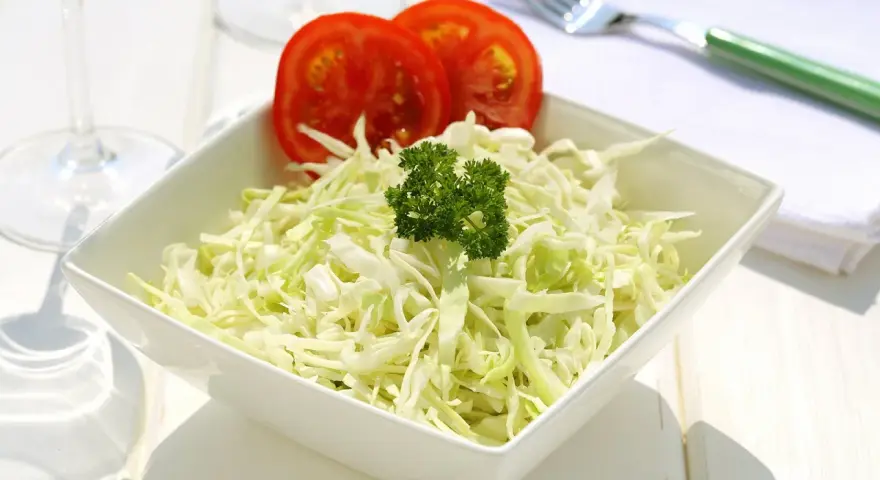 Frischer, selbstgemachter Krautsalat in einer weißen Schüssel, garniert mit Petersilie und Tomatenscheiben. Daneben liegen ein grüner Löffel und eine Serviette.