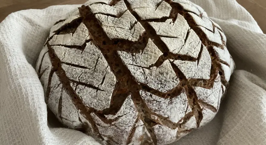 Ein rustikales, rundes buchweizenbrot sauerteig mit einer gemusterten Kruste, bestäubt mit Mehl, liegt auf einem weißen Leinentuch.