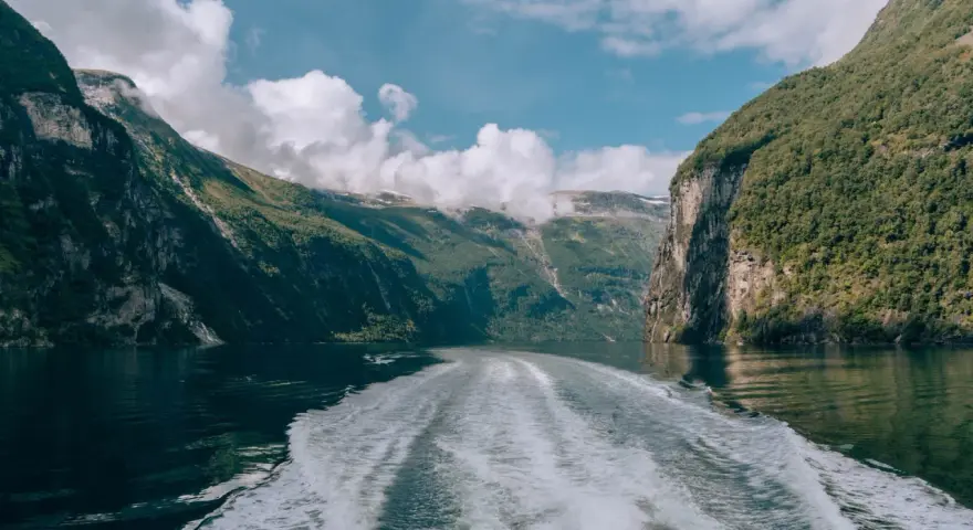 Rejs po Geirangerfjordzie. Pogoda sprzyja, chmury wiszą nad zielonymi zboczami gór, a za statkiem widać biały ślad na spokojnej wodzie.