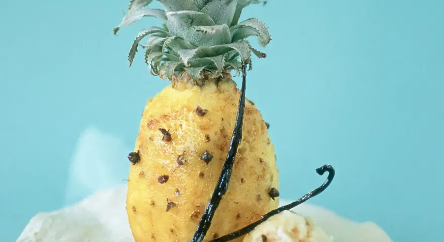 Un ananas rôti, parsemé de clous de girofle et de gousses de vanille, sur un fond bleu pâle. Un délice sucré et épicé.