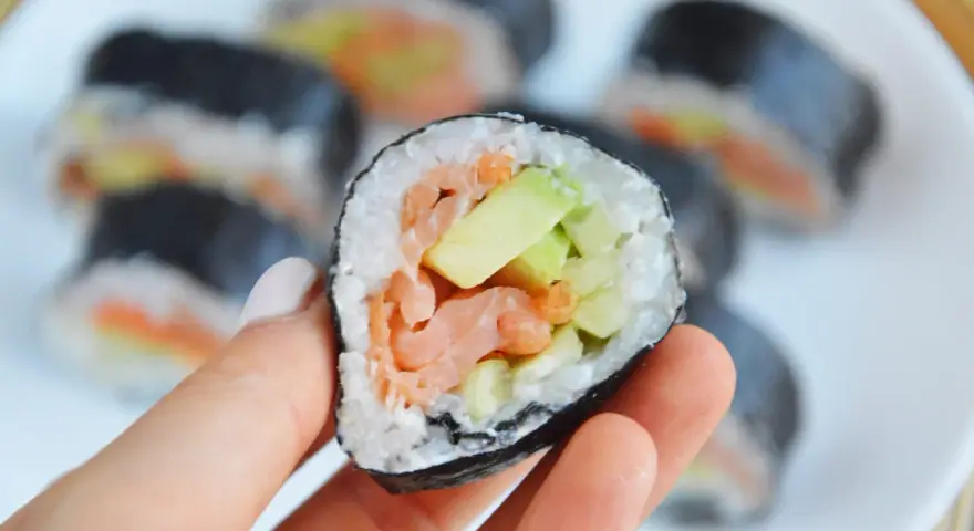 Palec wskazujący trzyma kawałek futomaki z łososiem, awokado i ogórkiem. Na talerzu widać więcej sushi.