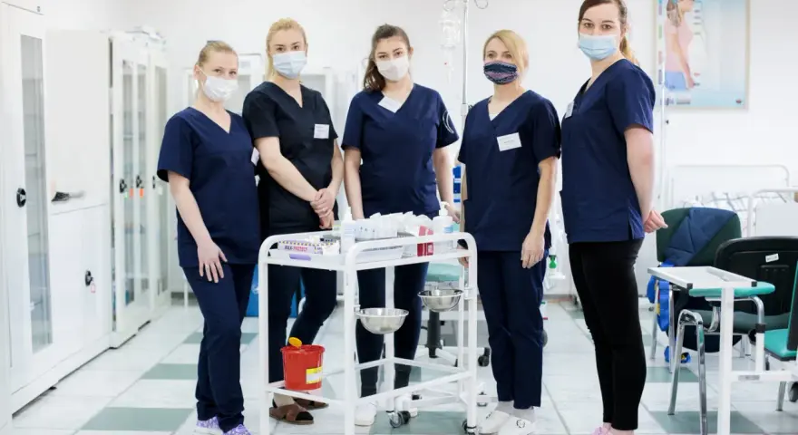 Pięć studentek pielęgniarstwa w granatowych uniformach i maseczkach stoi przy wózku medycznym. To ich przyszła praca dla studenta pielęgniarstwa.