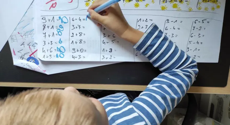 Dziecko rozwiązuje zadania matematyczne. Na kartce widać różne działania, a obok nich cyfry, które mogą stanowić **skalę ocen** lub odpowiedzi.