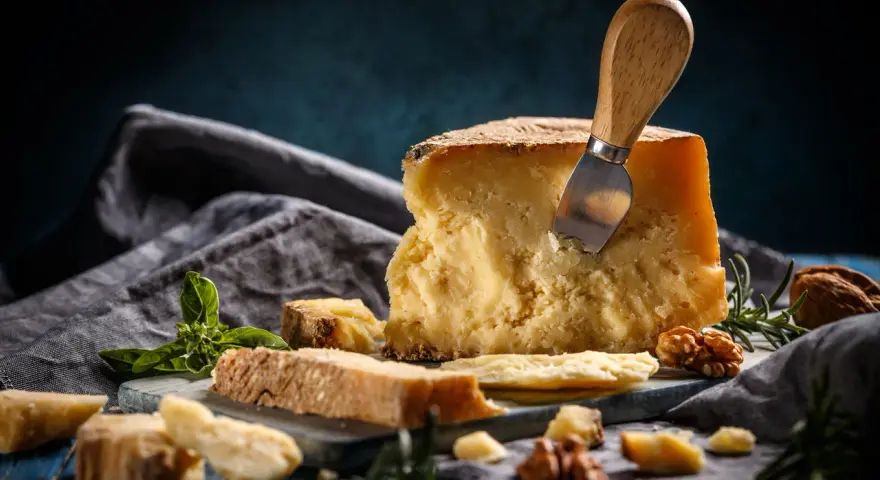Kawałek parmezanu z nożem, na desce z ziołami i orzechami.