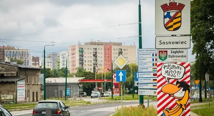 Znak drogowy "Sosnowiec" i humorystyczny napis "Paszport masz?!" przy wjeździe do miasta. W tle bloki mieszkalne.