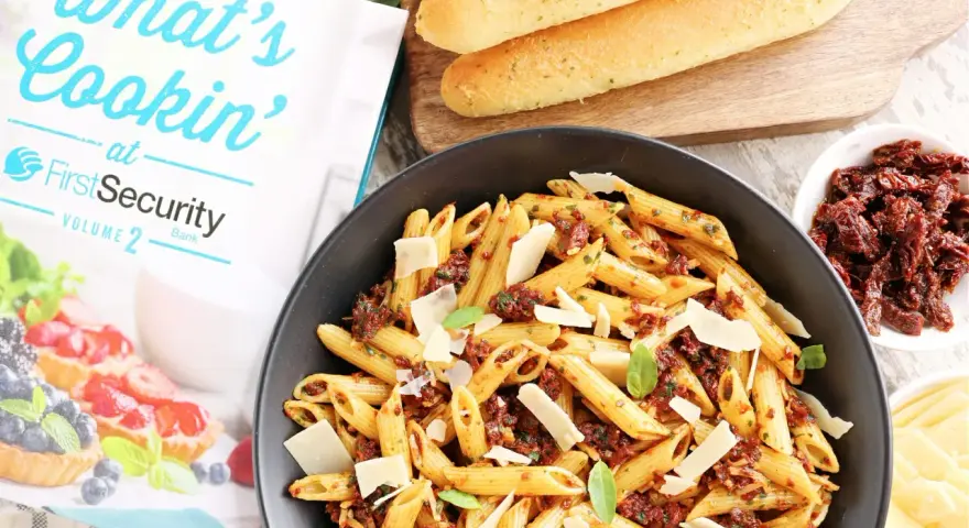 Makaron penne z suszonymi pomidorami, parmezanem i bazylią w czarnej misce, obok pieczywa i książki kucharskiej.