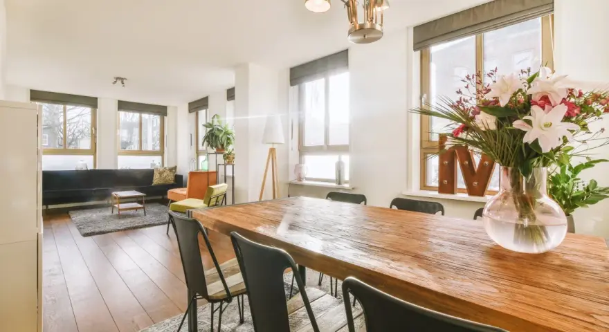 Amplio salón comedor dos ambientes con mesa de madera, sillas negras, sofá, sillones y flores.