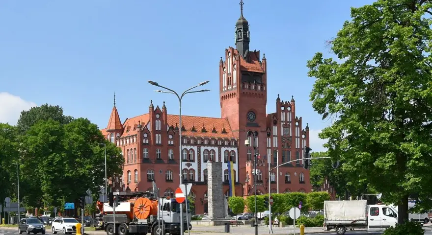 Imponujący ratusz w Słupsku, jedna z głównych atrakcji miasta, góruje nad placem. W tle zielone drzewa i błękitne niebo.