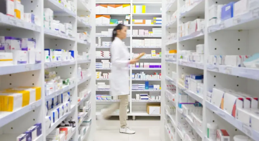 Farmaceutka w białym fartuchu idzie między regałami pełnymi leków, szukając amoksycyliny.