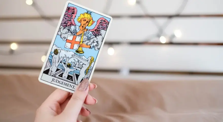 Una mano sostiene la carta de **el juicio tarot**, mostrando un ángel con trompeta y personas resucitando.