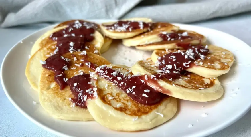 Placki serowe keto ułożone w okrąg na białym talerzu, polane konfiturą i posypane wiórkami kokosowymi.