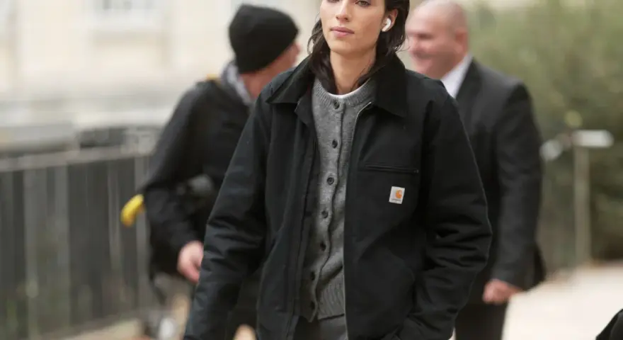 Donna con giacca Carhartt nera e cardigan grigio, cuffiette bianche, cammina con sicurezza.
