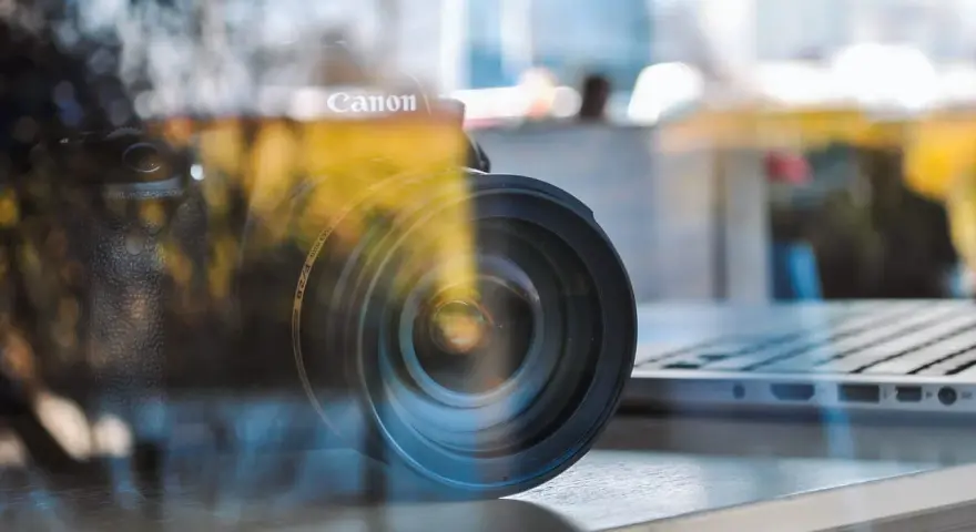 Winieta fotografia: obiektyw Canon na tle laptopa, odbicia tworzą artystyczny efekt.