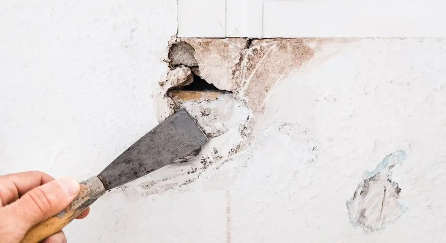 Mano con spatola rimuove intonaco da un buco nel muro troppo grande, rivelando mattoni e detriti.