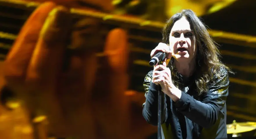 Ozzy Osbourne śpiewa do mikrofonu na koncercie, z rozmytym tłem w kolorach żółci i pomarańczy.