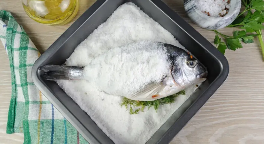 Dorada a la sal lista para hornear, con hierbas frescas y aceite de oliva al lado.