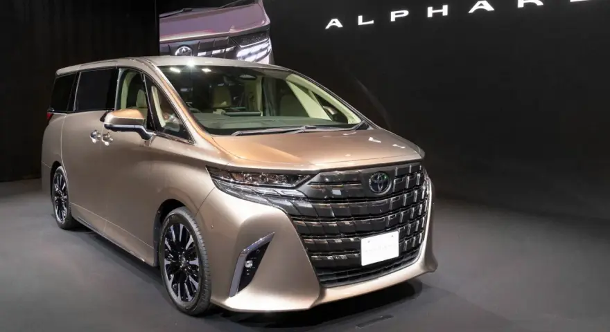 Brązowy minivan Toyota Alphard na ciemnym tle, z widocznym napisem "ALPHARD".