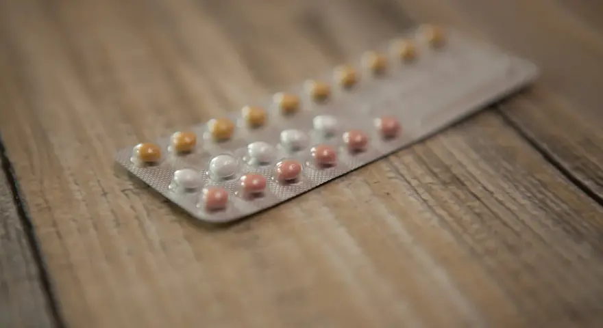 Opakowanie tabletek antykoncepcyjnych z białymi, różowymi i żółtymi pigułkami na drewnianym tle.