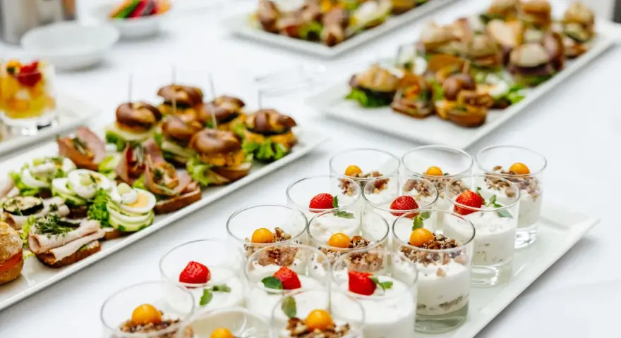 Varietà di antipasti freddi per buffet: mini hamburger, tartine con uova e salumi, e dolcetti in bicchiere con frutta e yogurt.