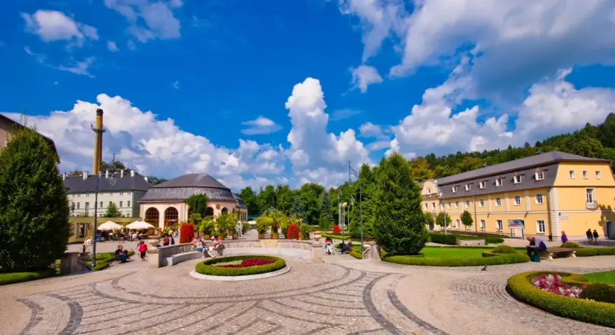Słoneczny dzień w Kudowie Zdrój. Spacerowicze odpoczywają w pięknym parku, podziwiając architekturę i zieleń. To idealne miejsce na relaks i odkrywanie atrakcji.