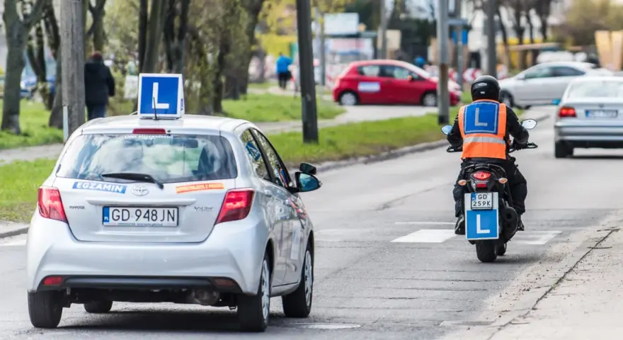 Srebrny samochód z literą "L" na dachu i motocyklista w kamizelce odblaskowej, obaj z literą "L" z tyłu, poruszają się po drodze. To kategoria a nauki jazdy.