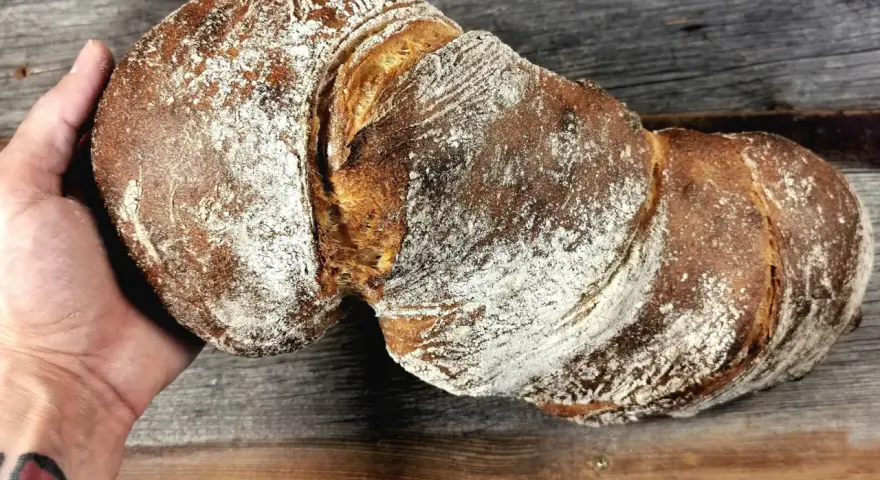 Ein rustikales wurzelbrot mit sauerteig, bestäubt mit Mehl, liegt auf einem Holzbrett.