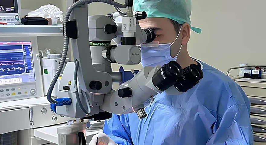 Chirurgo con maschera e camice blu opera con microscopio Zeiss. Monitor vitale e strumentazione indicano un intervento post-operatorio vitrectomia gas.