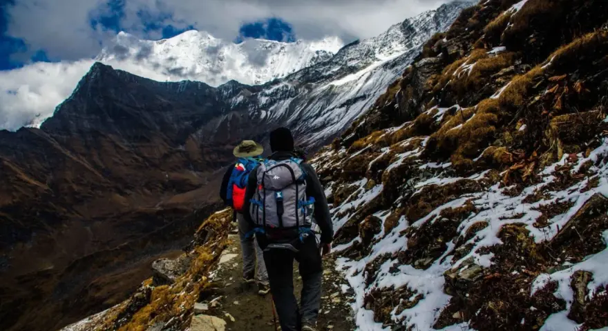 Dwóch turystów z plecakami wędruje ścieżką u podnóża ośnieżonych gór, przypominających krajobraz Mount Kenya.
