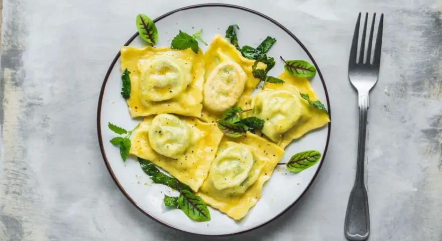 Pyszne ravioli z ricottą i szpinakiem, polane sosem, ozdobione świeżymi ziołami. Obok leży widelec.