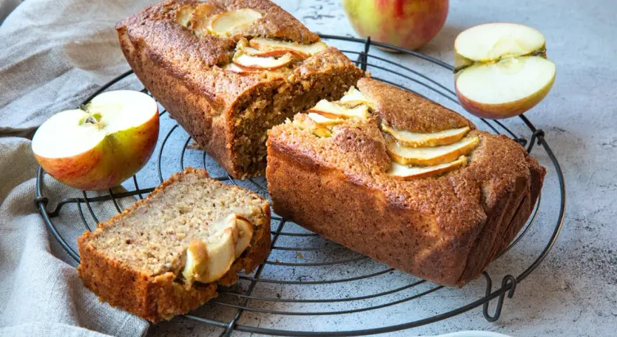 Saftiger Apfelkuchen mit Apfelmus-Note, dekoriert mit frischen Apfelscheiben. Daneben liegen Äpfel und ein Stück Kuchen.
