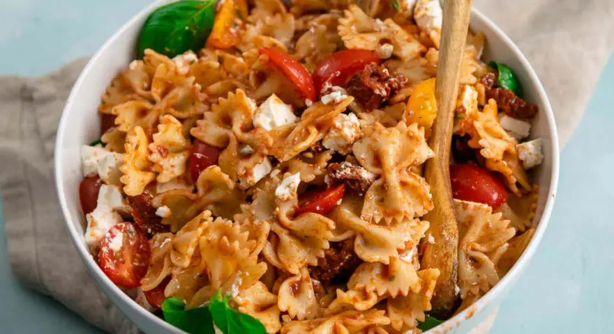 Ein köstlicher, sommerlicher Nudelsalat mit Farfalle, Tomaten, Feta und Basilikum, serviert in einer weißen Schüssel mit Holzutensilien.