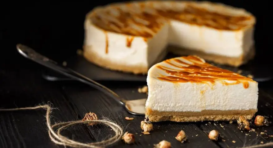 Una fetta di cheesecake senza panna, con glassa caramellata, su un piatto da portata, con la torta intera sullo sfondo.